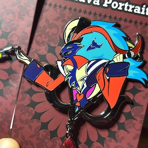 Peut inclure: &Eacute;pinglette color&eacute;e en &eacute;mail repr&eacute;sentant un personnage de "Helluva Boss". L'&eacute;pinglette, avec un contour noir, pr&eacute;sente un personnage aux accents bleus, oranges et jaunes. Un bijou rouge est suspendu en bas. La carte est rouge avec le texte "Helluva Portrait."