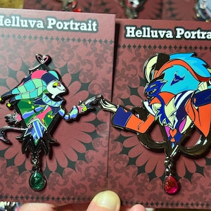 Peut inclure: Deux &eacute;pingles &eacute;maill&eacute;es repr&eacute;sentant des personnages de "Helluva Boss". L'une montre un personnage de type bouffon en vert, rose et bleu, l'autre un personnage orange, bleu et violet. Les deux &eacute;pingles ont une finition en m&eacute;tal fonc&eacute; et des pendentifs en pierres pr&eacute;cieuses.