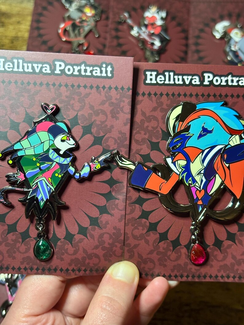 Peut inclure: Deux &eacute;pingles &eacute;maill&eacute;es repr&eacute;sentant des personnages de "Helluva Boss". L'une montre un personnage de type bouffon en vert, rose et bleu, l'autre un personnage orange, bleu et violet. Les deux &eacute;pingles ont une finition en m&eacute;tal fonc&eacute; et des pendentifs en pierres pr&eacute;cieuses.