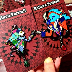 Puede incluir: Dos pines de esmalte con personajes de dibujos animados coloridos. Un pin representa a un bufón con una gema verde, el otro un personaje azul y naranja. Ambos pines están en tarjetas rojas con el texto "Helluva Portrait".