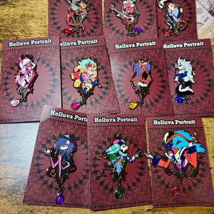 Peut inclure: Plusieurs &eacute;pingles &eacute;maill&eacute;es repr&eacute;sentant des personnages de "Helluva Boss". Chaque &eacute;pingle est fix&eacute;e sur une carte bordeaux avec le texte "Helluva Portrait". Les &eacute;pingles pr&eacute;sentent divers personnages color&eacute;s avec des motifs complexes et des accents de pierres pr&eacute;cieuses pendantes.