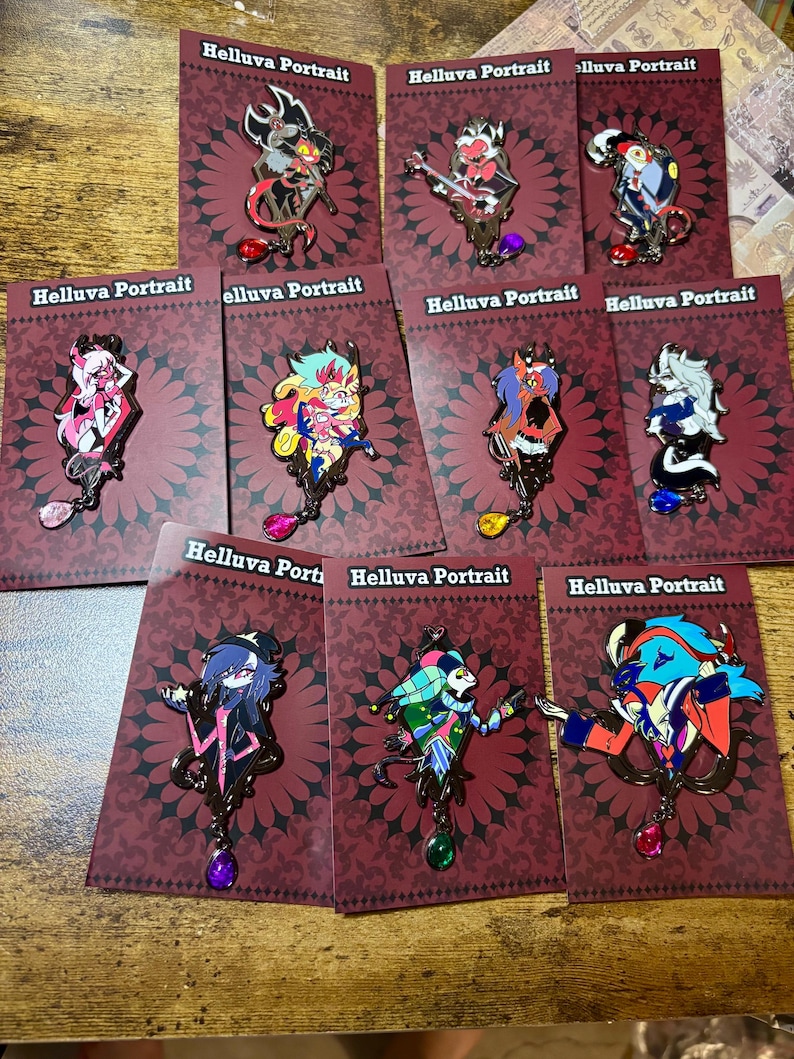 Peut inclure: Plusieurs &eacute;pingles &eacute;maill&eacute;es repr&eacute;sentant des personnages de "Helluva Boss". Chaque &eacute;pingle est fix&eacute;e sur une carte bordeaux avec le texte "Helluva Portrait". Les &eacute;pingles pr&eacute;sentent divers personnages color&eacute;s avec des motifs complexes et des accents de pierres pr&eacute;cieuses pendantes.