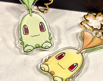 May include: Two acrylic keychains featuring a cartoon character with a green body, red eyes, and a leaf-like head. One keychain is green, the other is yellow. Both have gold-colored key rings.