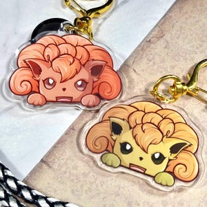 May include: Two clear acrylic keychains featuring a cartoon illustration of a Pokemon character with curly orange hair. The character is peeking over the top of the keychain. The keychains are attached to gold-colored metal key rings.