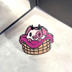 May include: A pink pig with black eyes and a black nose is nestled in a brown wicker basket with a pink blanket. The pig is wearing a pink blanket and is looking at the camera.