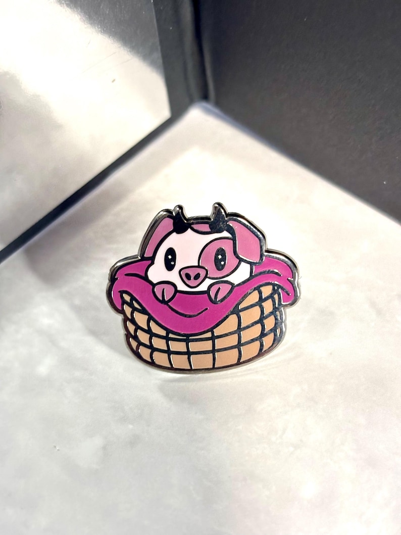 May include: A pink pig with black eyes and a black nose is nestled in a brown wicker basket with a pink blanket. The pig is wearing a pink blanket and is looking at the camera.