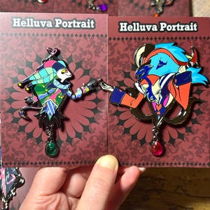 Peut inclure: &Eacute;pingles en &eacute;mail repr&eacute;sentant des personnages de Helluva Boss, pr&eacute;sent&eacute;es sur des cartes rouges avec le texte "Helluva Portrait". Les &eacute;pingles pr&eacute;sentent des motifs d&eacute;taill&eacute;s aux couleurs vives, dont le bleu, le rouge, le vert et le violet. Chaque &eacute;pingle a un bijou pendant.