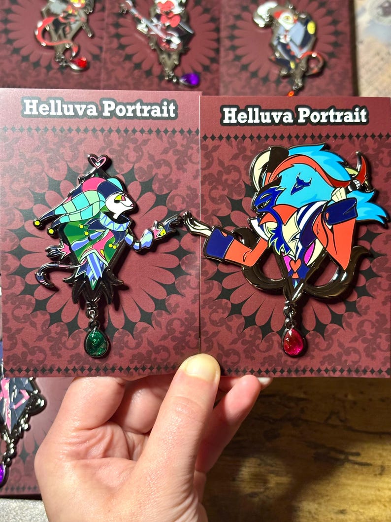 Peut inclure: &Eacute;pingles en &eacute;mail repr&eacute;sentant des personnages de Helluva Boss, pr&eacute;sent&eacute;es sur des cartes rouges avec le texte "Helluva Portrait". Les &eacute;pingles pr&eacute;sentent des motifs d&eacute;taill&eacute;s aux couleurs vives, dont le bleu, le rouge, le vert et le violet. Chaque &eacute;pingle a un bijou pendant.