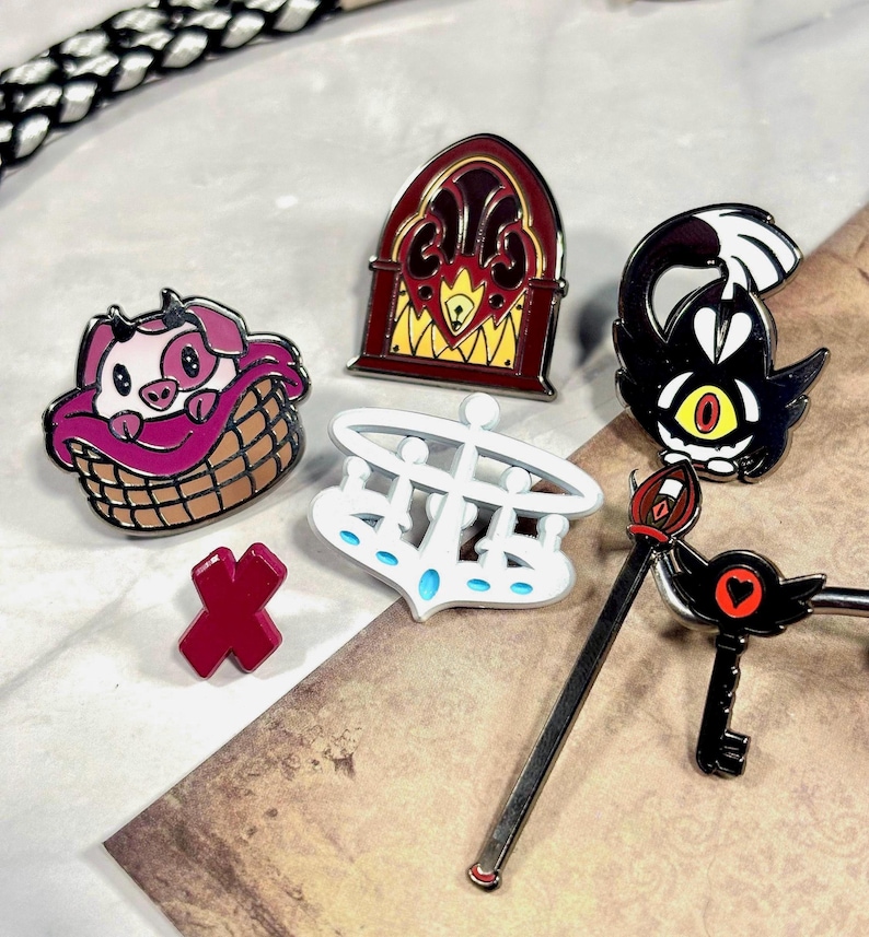 May include: A set of six enamel pins featuring characters and objects from the popular cartoon series "The Owl House". The pins include a pig in a basket, a radio, a crown, a key, a wand, and a demon with a single eye.