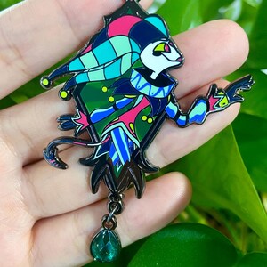 Peut inclure: &Eacute;pingle &eacute;maill&eacute;e color&eacute;e repr&eacute;sentant un personnage de bouffon avec un corps en forme de losange. L'&eacute;pingle a un contour noir et est orn&eacute;e d'accents roses, bleus, verts et jaunes. Un charme vert en forme de goutte d'eau est suspendu en bas.