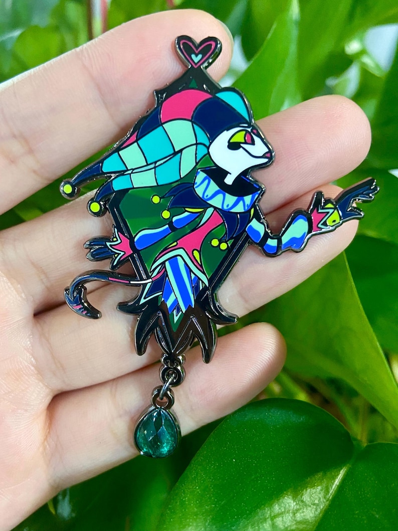 Peut inclure: &Eacute;pingle &eacute;maill&eacute;e color&eacute;e repr&eacute;sentant un personnage de bouffon avec un corps en forme de losange. L'&eacute;pingle a un contour noir et est orn&eacute;e d'accents roses, bleus, verts et jaunes. Un charme vert en forme de goutte d'eau est suspendu en bas.