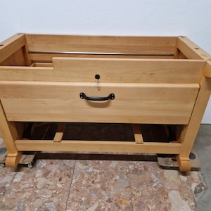 Könnte beinhalten: Ein helles Holzmöbel mit einer Schublade und einem schwarzen Griff. Der Schrank hat eine rechteckige Form mit abgerundeten Kanten und steht auf kleinen Rollen. Über die Oberseite verläuft eine Metallstange.
