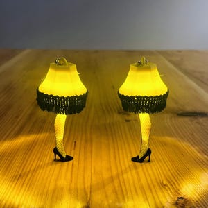 May include: Two novelty lamps shaped like a woman's leg in a fishnet stocking and high heel. The lamps have a yellow lampshade with black fringe. The lamps are on a wooden surface.