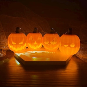 May include: Four glowing orange pumpkin decorations with carved faces, lit from within, sit on a light-colored platform. The Halloween-themed pumpkins have varying expressions and are set against a dark background.