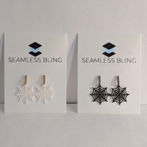 May include: Two pairs of spiderweb earrings on white cards. One pair is white and the other is black. The cards have the text "Seamless Bling" on them.