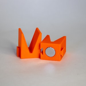 May include: Two orange plastic 3D printed objects. One object is a V-shaped stand with a flat base. The other object is a cube with a round, silver magnet on one side.