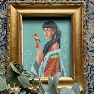 May include: A framed art print featuring a woman with dark hair and a colorful bird perched on her finger. The artwork has a teal background with floral accents and is set in a gold ornate frame. The woman is wearing an orange and teal garment.