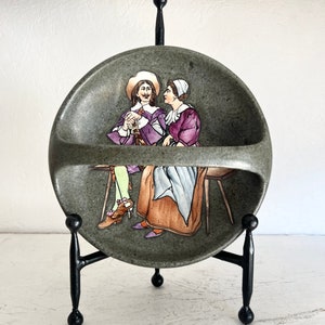 May include: A ceramic wall pocket with a gray background and a painted scene of two people in period clothing. The pocket has a cutout for hanging on a wall.