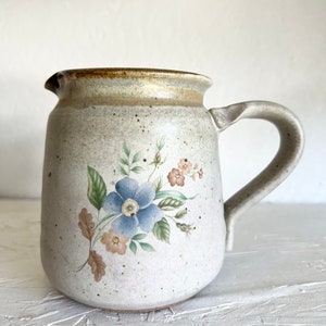 May include: A white ceramic pitcher with a brown rim and a floral design featuring blue and pink flowers. The pitcher has a handle and a speckled glaze.