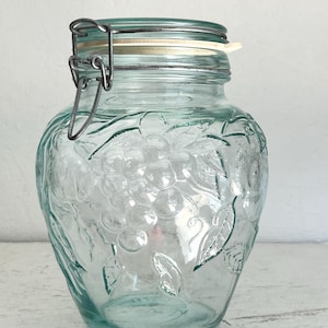 May include: A clear glass jar with a metal lid and a grapevine design. The jar has a light green tint and is perfect for storing food.