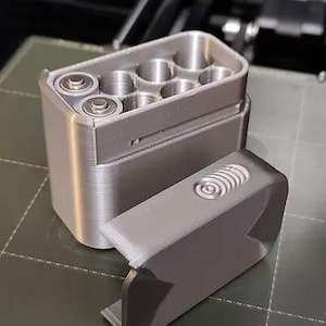 May include: A silver-colored, 3D printed battery storage case with a hinged lid. The case has six compartments for storing batteries.