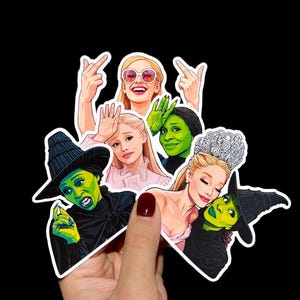 Puede incluir: Una colección de pegatinas con ilustraciones de personajes del musical "Wicked". Las pegatinas representan a los personajes en varias poses, con colores vibrantes y contornos audaces. Las pegatinas se sostienen sobre un fondo negro.