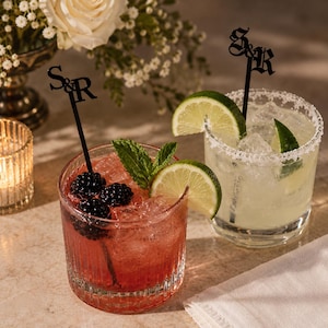 May include: Two black acrylic drink stirrers featuring the letters S&R in a gothic font. One stirrer is in a red cocktail with blackberries and mint, and the other is in a clear cocktail with a salted rim and lime wedges in glass tumblers.