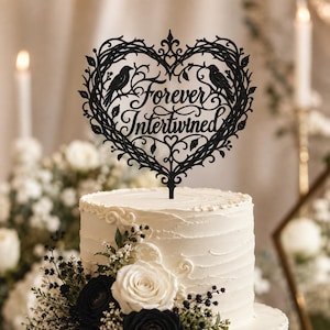 May include: A black cake topper featuring a heart-shaped frame made of intricate vines and leaves. Two birds are perched on the branches within the heart. The text "Forever Intertwined" is written in a cursive script in the center of the heart.