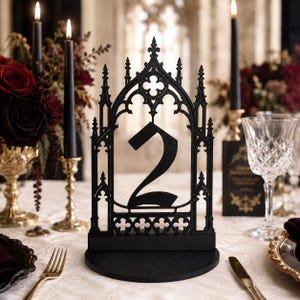 May include: A black table number sign featuring the number 2 centred within an intricate, pointed arch frame inspired by gothic architecture. The sign is mounted on a circular base and is constructed with detailed cutouts and finials.
