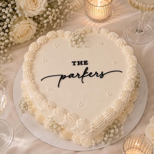 May include: A heart-shaped cake with smooth white frosting, decorated with piped borders, small white pearl accents, and sprigs of baby's breath. The top features the black text "THE parkers" written in a combination of block and script fonts.