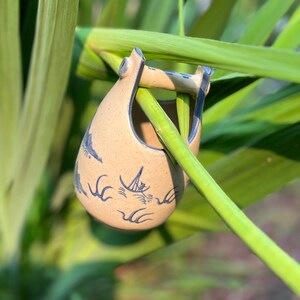 May include: A small, beige ceramic planter with a blue and white design of a sailboat and waves. The planter is hanging from a green plant.