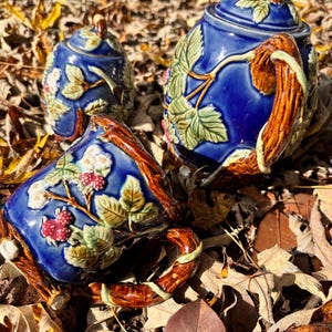 May include: A blue ceramic tea set with a teapot, sugar bowl, and teacup. The set features a raised design of leaves and berries, with brown handles and accents. The set is sitting on a bed of fallen leaves.