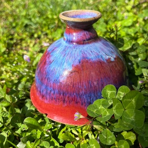 May include: A small, red and blue ceramic vase with a wide, rounded base and a narrow neck. The vase is sitting on a bed of green clover.