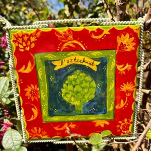 May include: A red and green square tile with a yellow border and a green artichoke illustration in the center. The tile has the text "l'artichaut" in a yellow banner above the artichoke.