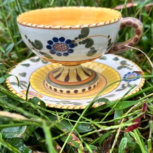 May include: A hand-painted ceramic teacup and saucer set. The cup and saucer feature a white base with floral and leaf designs in blue and green. The cup's interior and the saucer's center are yellow. The handle has a brown speckled pattern.