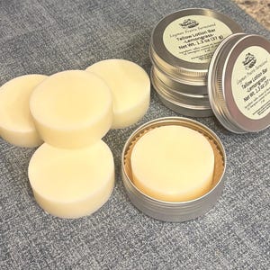 May include: A collection of round, pale yellow tallow lotion bars, some stacked and others in small, silver tins. The tins have labels with text that reads "Layman Prairie Farmsand Tallow Lotion Bar - Lemongrass - Net Wt. 1.3 oz (37g)".