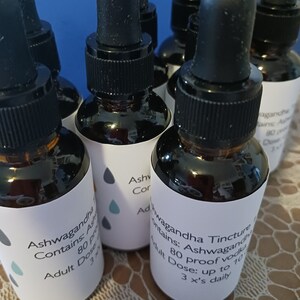 May include: A close-up of five amber glass bottles with dropper tops. The bottles are labeled with white labels that say "Ashwagandha Tincture" and "Contains: Ashwagandha 80 Proof Vodka Adult Dose: up to 10 drops 3 x's daily".
