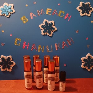 May include: Eight small brown glass bottles with gold lids and labels with a tree design. The bottles are arranged in a row on a wooden surface. The background is a blue wall with the words "Sameach Chanukah" in colorful letters.