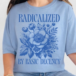 May include: A light blue t-shirt with the words "RADICALIZED" and "BY BASIC DECENCY" in blue text. A blue floral design is centered below the word "RADICALIZED". The shirt is made of a soft material and has short sleeves.