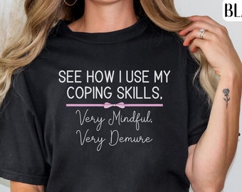 May include: Black t-shirt with the text "See how I use my coping skills. Very mindful. Very demure." in white text with a pink bow above the text.