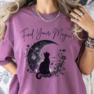 May include: A purple t-shirt with the text "Find Your Magic" and a black silhouette of a cat sitting on a crescent moon with floral accents.