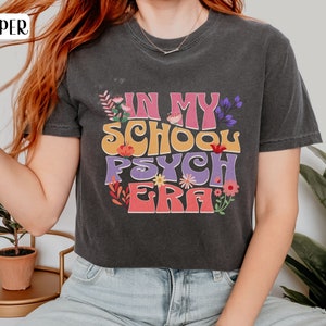 May include: A dark gray cropped t-shirt with the text "In My School Psych Era" in colorful, retro-style lettering. The text is surrounded by small floral designs.