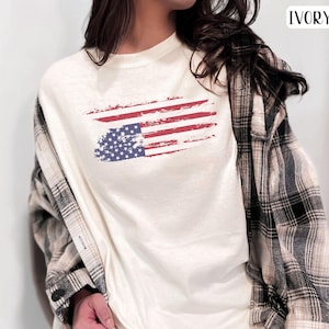 May include: A white t-shirt with a distressed American flag design printed on the front. The shirt is being worn by a model and has a relaxed fit.