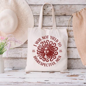 May include: Cream-colored tote bag with a red Medusa illustration and the text "I WILL NOT YIELD TO DISRESPECTFUL MEN". The bag has two handles and is made of a sturdy fabric. A straw hat and a beige drawstring bag are in the background.