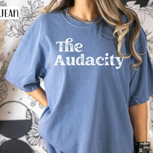 Puede incluir: Una camiseta de mezclilla azul con el texto "The Audacity" impreso en blanco en la parte delantera.