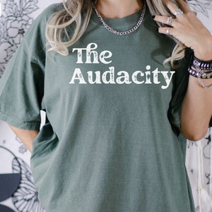 Puede incluir: Una camiseta verde con el texto "The Audacity" impreso en blanco en la parte delantera.