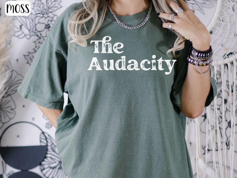 Puede incluir: Una camiseta verde con el texto "The Audacity" impreso en blanco en la parte delantera.