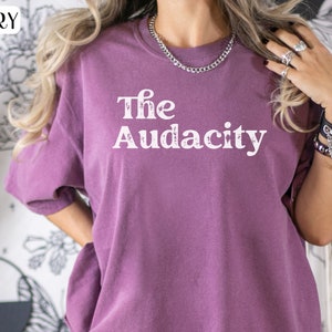 Puede incluir: Una camiseta morada con el texto "The Audacity" impreso en blanco en la parte delantera.