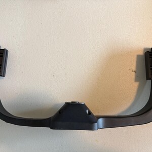May include: A matte black, U-shaped plastic bracket featuring a central mounting point with two screw holes and two vertical side attachments. The component has a smooth, curved frame design with reinforced edges and a functional, industrial appearance.