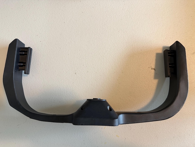 May include: A matte black, U-shaped plastic bracket featuring a central mounting point with two screw holes and two vertical side attachments. The component has a smooth, curved frame design with reinforced edges and a functional, industrial appearance.